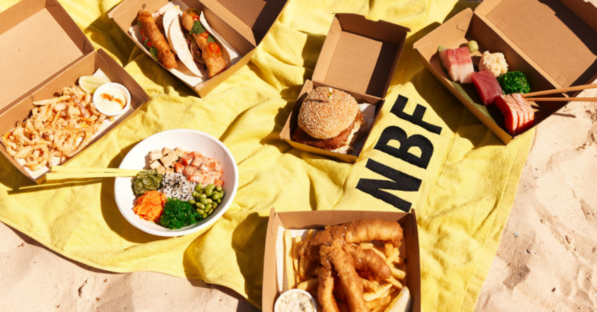 Selection of North Bondi Fish takeaway dishes including fish tacos, burger, poke bowl and battered fish on a yellow beach towel.