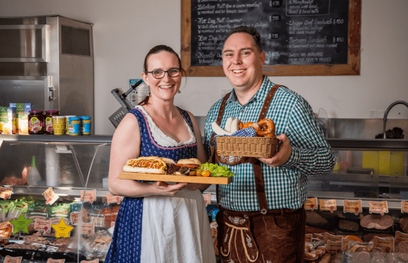 Man and Woman in German costume holding pretzels and sausages at butcher counter of a German Butchery in Bexley North with meat in the background,