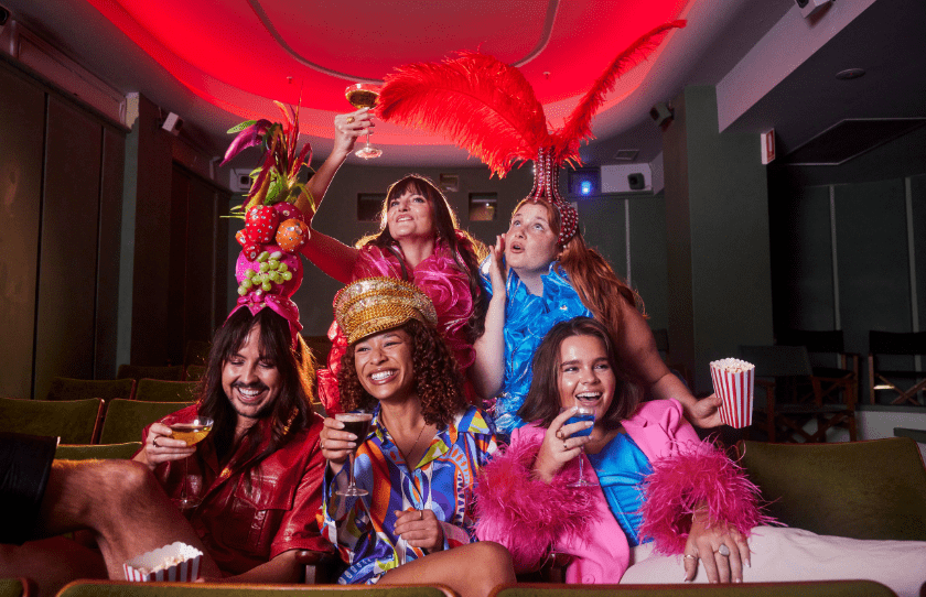 Drag queen and girls dressed up in 1994 costumes with sequins and head pieces holding popcorn, martinis and sitting in the Golden Age Cinema 
