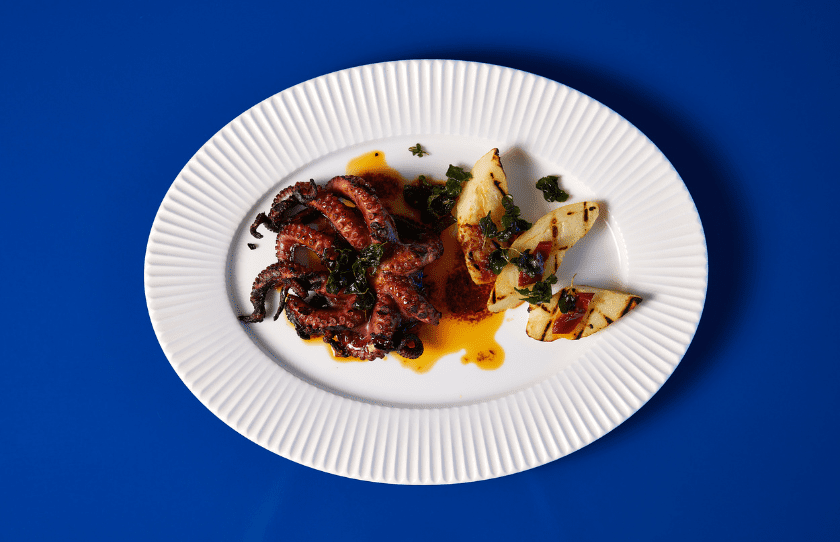 WA octopus brined in a fragrant mix of Vermentino, garlic and citrus served with kipler potatoes on a white plate with a blue background